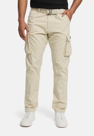 Man draagt beige cargobroek met meerdere zakken, een beige riem, witte sneakers en een zwart shirt, staand met handen dicht bij de zakken.