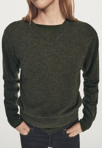 Personne portant un pull à col en V vert foncé superposé sur une chemise verte, les mains dans les poches avant d'un jean bleu foncé.