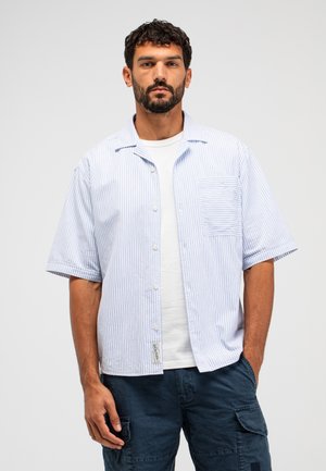 Man met baard, gekleed in een lichtblauw gestreept shirt met korte mouwen over een wit shirt en donkere cargobroek, staand tegen een effen achtergrond.
