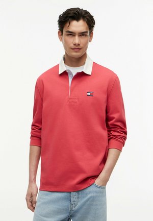 Junger Mann mit dunklen Haaren, der ein korallenfarbenes Langarm-Poloshirt mit weißem Kragen und hellblauen Jeans trägt, steht vor weißem Hintergrund.