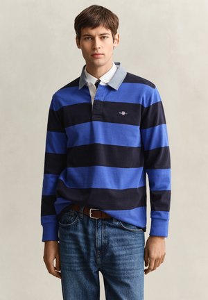 Junger Mann trägt ein langärmeliges Rugbyhemd mit blauen und schwarzen horizontalen Streifen und blaue Jeans, steht vor einem einfarbigen Hintergrund.