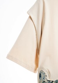 Camisa de manga corta beige con costuras visibles sobre un fondo blanco, mostrando parte de una tela de patrón azul y gris debajo.