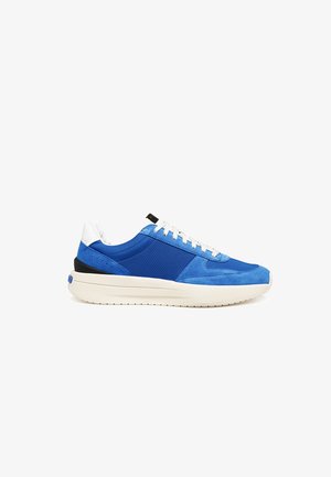 Blauwe en witte lage sneaker met bovenwerk van suède en mesh, witte veters en dikke witte zool tegen een witte achtergrond.