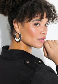 Black suede jacket with silver hardware details, featuring a high collar and button accents. Model displays large silver hoop earrings.