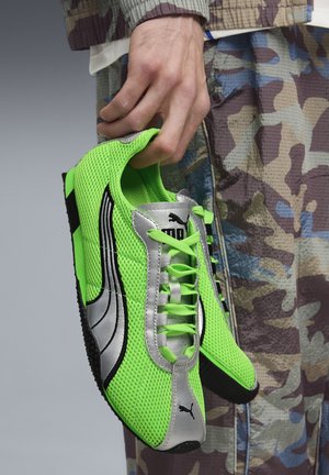 Hand holding fluorescent green Puma sneakers with silver and black details, wearing camouflage-patterned pants.