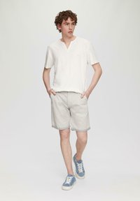 Witte korte mouwen, v-hals shirt gecombineerd met lichtgrijze shorts. Het model draagt blauwe sneakers met witte accenten. Eenvoudige, casual outfit.