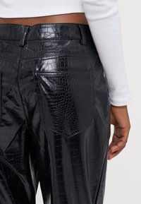 Närbild på en person som bär glänsande svarta byxor i imitationskrokodilläder och en vit långärmad topp, där bakfickan och handen syns.