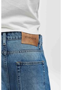 Jeans i denim med en ljus tvätt, med en brun läderetikett präglad med "2Y STUDIOS" baktill, femficksdesign och sydda detaljer.