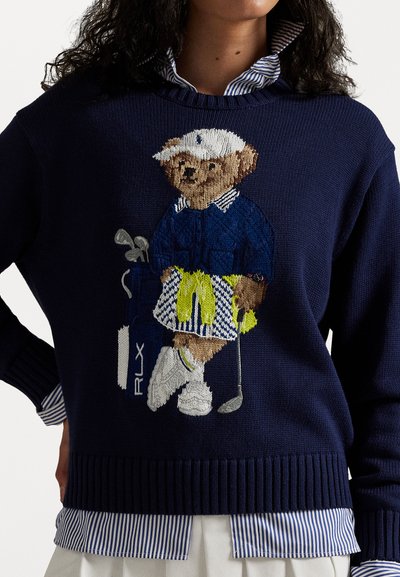 Navy knitted sweater featuring an embroidered bear in golf attire, holding a club with accent colors in yellow and white. Striped collar visible.