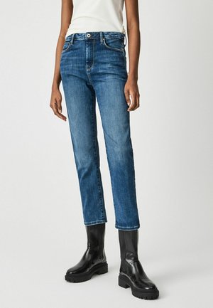 Persoon die blauwe rechte jeans en zwarte stevige leren laarzen draagt, staand tegen een effen lichte achtergrond.