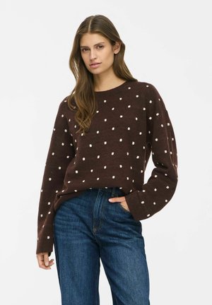 Une femme porte un pull marron foncé à pois blancs et un jean bleu. Elle se tient debout, une main dans la poche, devant un fond blanc uni.