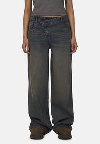 Jeans en denim à jambe large de couleur bleu foncé avec un effet délavé, dotés d'une fermeture à bouton et de poches avant. Assortis à un haut gris ajusté et des chaussures marron.