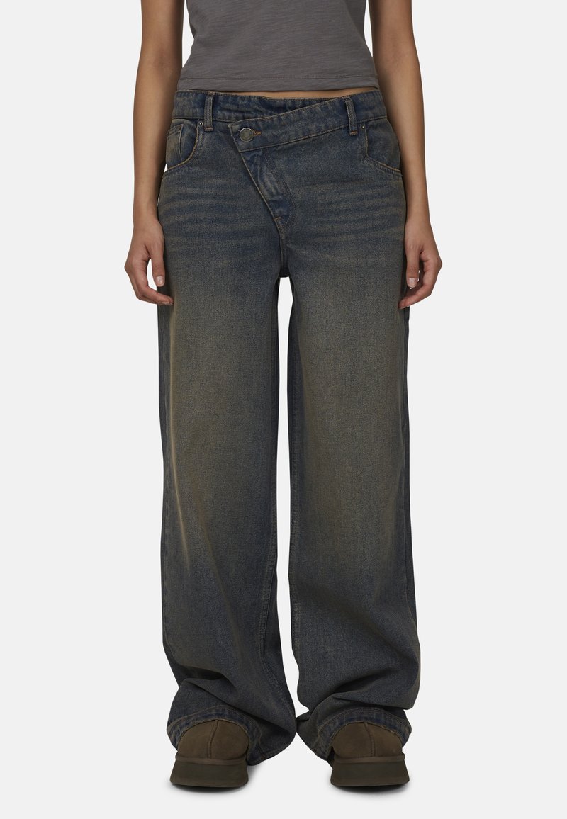 Jeans en denim à jambe large de couleur bleu foncé avec un effet délavé, dotés d'une fermeture à bouton et de poches avant. Assortis à un haut gris ajusté et des chaussures marron.