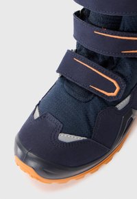 Jūras zila zābaki ar velcro siksnām, izgatavoti no tekstila un gumijas materiāliem, ar pelēkiem akcentiem un oranžu zoli. Iezīmējas ar teksturētu auduma dizainu.