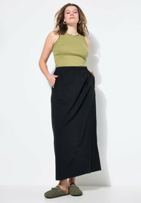 Olijfgroen geribbelde tanktop met geschulpte randen, gecombineerd met een lange zwarte rok met zijzakken. Model draagt groene instapschoenen.