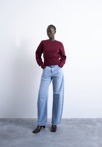 Femme debout les mains dans les poches, portant un pull bordeaux, un jean large bleu clair et des bottes à talons foncées sur un sol gris.