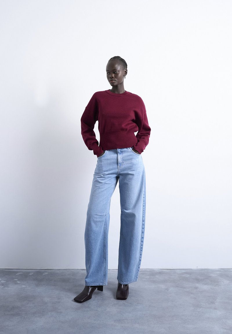 Femme debout les mains dans les poches, portant un pull bordeaux, un jean large bleu clair et des bottes à talons foncées sur un sol gris.