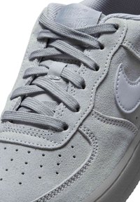 Světlé šedé semišové tenisky s texturovaným povrchem, vybavené plochými šedými tkaničkami, malými perforacemi na špičce a bílým logem jako akcentem.