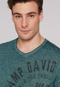 Camiseta a rayas verdes con un diseño de cuello en V, que presenta un estampado gráfico oscuro con la inscripción "AMP DAVID" y texto abajo, fabricada con una tela texturizada.