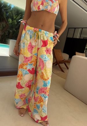 Naine kannab mitmevärvilisi tikandihallisid pükse ja sobivat lühikest tuba, seisab paljajalu siseruumides puidust toolide ja beeži diivani lähedal.