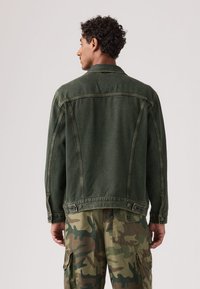 Χακί τζιν μπουφάν με άνετη γραμμή, ορατές ραφές και λεπτομέρειες με κουμπιά στη μέση, συνδυασμένο με παραλλαγμένα cargo παντελόνια.