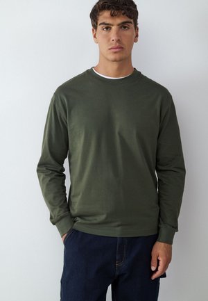 Young man with short dark curly hair wears olive green long-sleeve shirt and dark blue jeans standing against a plain white background.