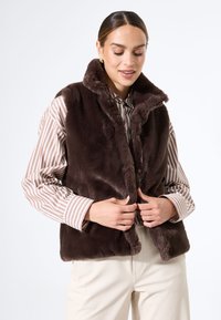 Braune Kunstfellweste mit hohem Kragen, die eine glatte Textur und einen Frontverschluss aufweist, getragen über einem gestreiften Hemd mit Knöpfen.