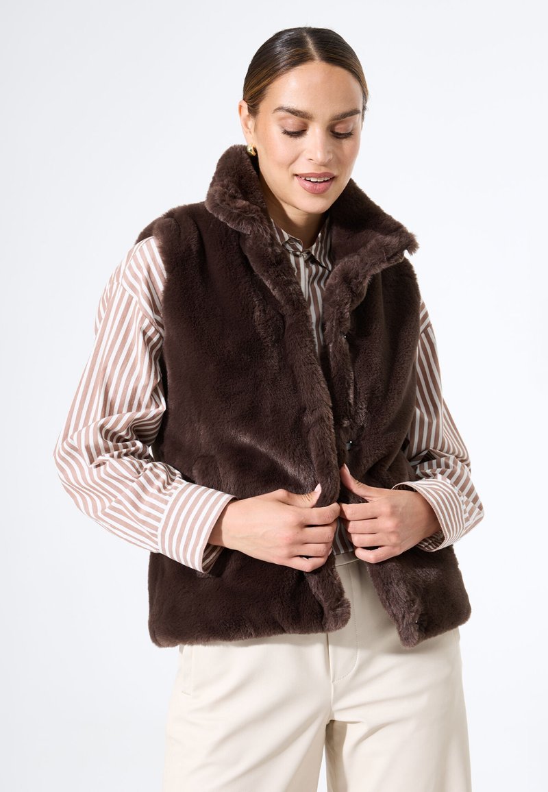 Braune Kunstfellweste mit hohem Kragen, die eine glatte Textur und einen Frontverschluss aufweist, getragen über einem gestreiften Hemd mit Knöpfen.