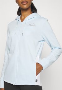 Femme portant un sweat à capuche zippé Puma bleu clair avec capuche et cordons, main droite dans la poche, vue de la nuque aux hanches.