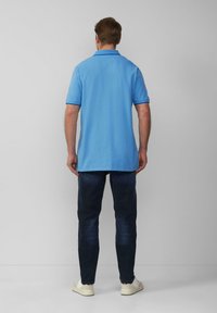 Man in een lichtblauw poloshirt met korte mouwen, donkere jeans en witte sneakers staand voor een effen lichtmuur.