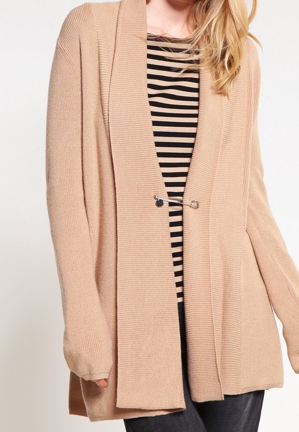 Femme portant un cardigan beige ouvert devant, maintenu par une épingle de sûreté, sur un haut rayé noir et beige et un pantalon foncé.