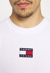 Camiseta de algodón blanca con un parche bordado rectangular que dice "TOMMY JEANS" y detalles en color rojo y blanco. Cuello redondo.
