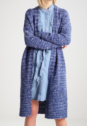 Cardigan blu testurizzato con maniche lunghe, apertura frontale e design a maglia indossato sopra una camicia di jeans azzurra chiara.