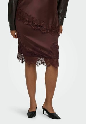 Jupe en satin rouge foncé avec bordure en dentelle portée avec des chaussures noires à talons hauts et slip-on et une veste en cuir noire visible sur les bras.