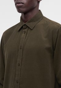 Gros plan d'une personne portant une chemise en velours côtelé vert olive boutonnée avec un col pointu sur un fond uni.