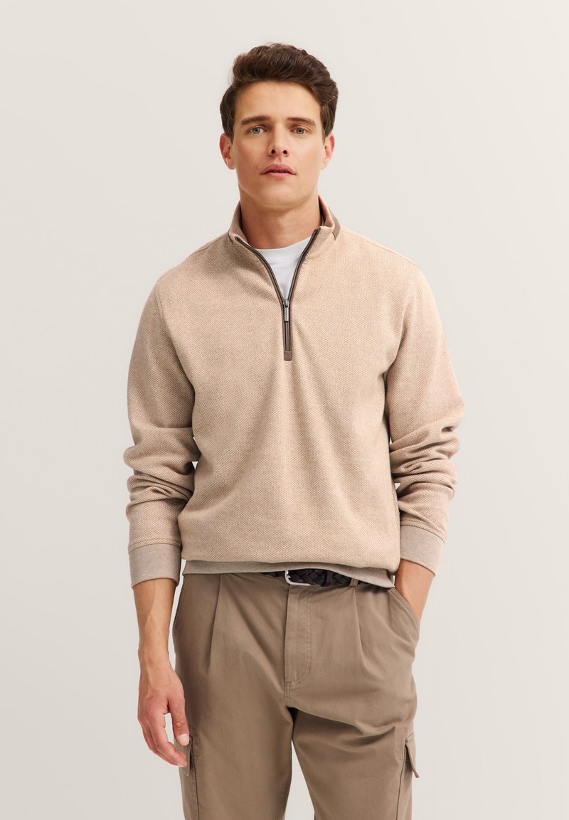 Beige Zip-Up-Pullover mit strukturiertem Finish, gerippten Bündchen und Stehkragen. Kombiniert mit hellbraunen Hosen.