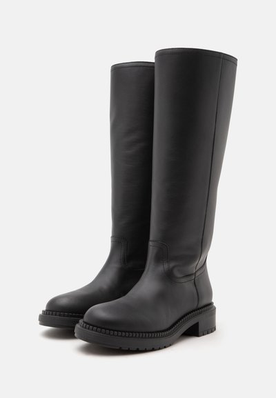 Paire de bottes en cuir noires montantes jusqu'aux genoux avec semelles épaisses et talons bas sur fond blanc.