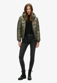 Giacca puffer verde scuro con cappuccio, caratterizzata da una texture trapuntata e un logo rosso, indossata sopra un maglione nero e jeans skinny con stivali militari.