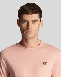 Homme avec les cheveux bruns courts et les yeux bleus portant un t-shirt à col rond rose clair avec un petit logo d'oiseau jaune sur la poitrine.