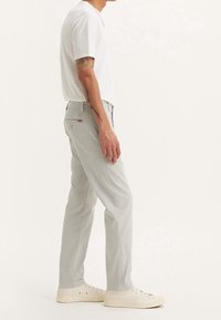 Jasnoszare spodnie proste wykonane z bawełnianego płótna, z tylną kieszenią z naszywką z logo oraz subtelnymi detalami szycia. Noszone z białą koszulą.