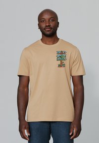 Beige T-shirt met korte mouwen en een kleurrijke grafische print van richtingaanwijzers op de linkerkant van de borst. Gemaakt van zacht katoen.