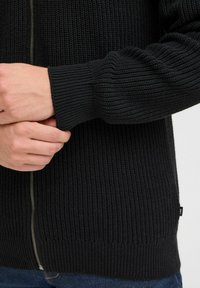 Svart strikket cardigan med ribbet tekstur, som har glidelås foran og tettsittende ermer. Bunnkanten har ribbet hem og en liten merkelapp.
