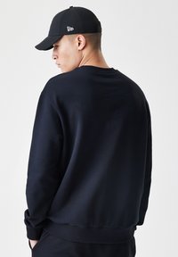 Sort sweatshirt med rund hals og ribbede manchetter. Stoffet ser glat og tykt ud. En sort baseballcap bæres også, med et logo.