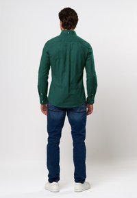 Zöld, hosszú ujjú ing, gombos gallérral, hátsó redőzéssel, kék farmernadrággal és fehér tornacipővel párosítva, hátulról nézve.