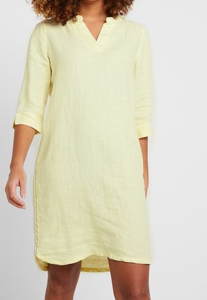 Femme portant une robe en lin ample, longueur genou, jaune pâle, avec des manches trois-quarts et une petite découpe en V au niveau du col.