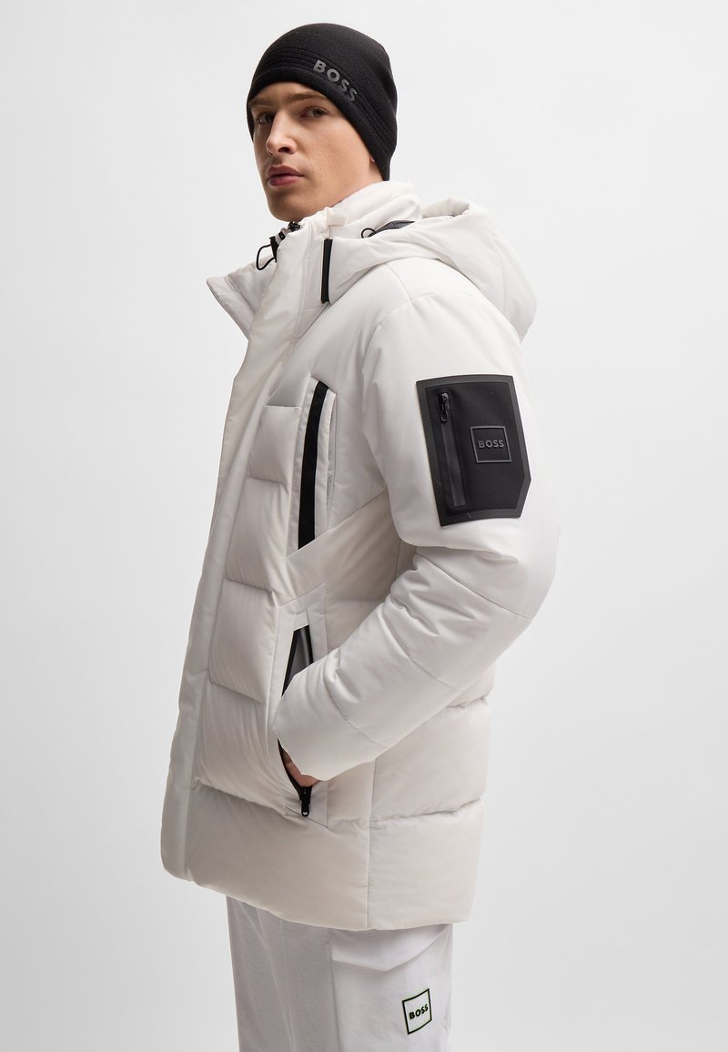 Gepolsterte weiße Jacke mit Kapuze, schwarzen Akzenten, Reißverschlusstaschen und einem Markenpatch auf dem linken Ärmel. Getragen mit einer schwarzen Mütze.