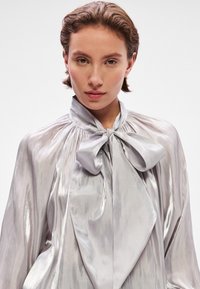 Silvery blouse with a high collar and front bow tie detail. The fabric is shiny, featuring a smooth texture and subtle draping.
