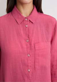 Różowa lniana koszula z kołnierzykiem na guziki, z guzikami z przodu i kieszonką na piersi. Tkanina ma lekko teksturowany wygląd.