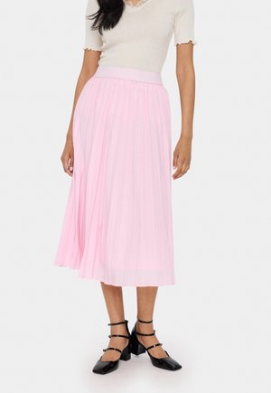 Mujer con un top crema de manga corta, falda midi plisada rosa claro y sandalias negras de tiras, de pie contra un fondo liso.