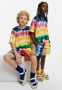 Due bambini indossano t-shirt e pantaloni corti abbinati di colore vivace, con fantasie di blu, rosso, giallo e verde. Ai piedi indossano sandali.